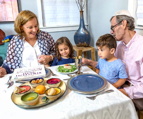 Jewish grandparents and two young children gathered around a dinner table with a seder plate and kiddush cup, celebrating Passover.